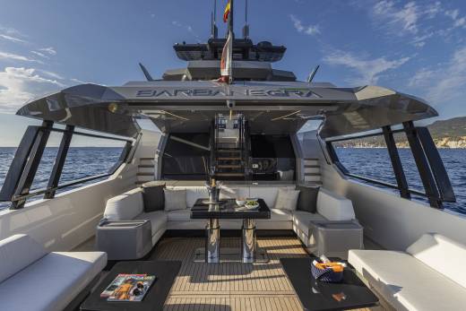 A luxurious yacht deck with plush seating and tables in the foreground is set against a backdrop of the open sea and a distant coastline under a clear blue sky.
