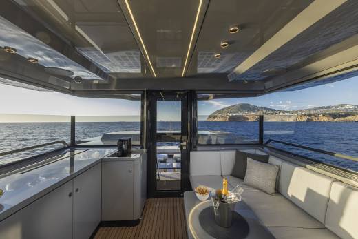 A modern yacht's interior lounge with a wet bar in the foreground and a scenic ocean view with a hilly coastline in the background.
