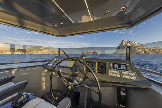 A modern yacht's helm with a large steering wheel and digital displays is positioned in the foreground, overlooking a calm sea and a rocky island in the background under a clear sky.
