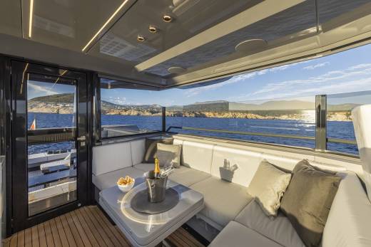 A luxurious yacht's outdoor seating area with a table holding champagne and snacks is set against a backdrop of a calm blue sea and a distant, sun-drenched coastline.