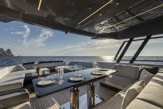 A luxurious yacht's outdoor dining area with a table set for a meal is in the foreground, while the calm ocean and a distant island are visible in the background under a bright sky.