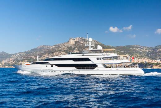 A large white yacht cruises on blue water in the foreground, with a mountainous coastline dotted with buildings in the background under a clear blue sky.