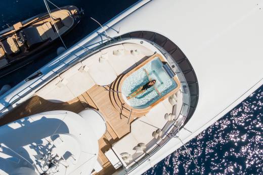 A woman relaxes in a jacuzzi on the deck of a large yacht, with a smaller boat docked nearby and sparkling blue water in the background.