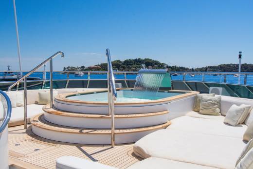 A circular jacuzzi with a waterfall feature is in the foreground, while the background shows a calm blue sea dotted with boats and a lush green coastline under a clear sky.