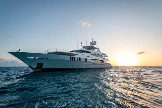 A large white yacht floats on calm blue water in the foreground, with a golden sunset visible on the horizon in the background.