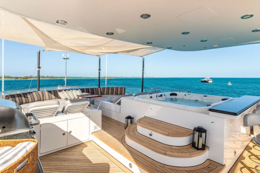 A luxurious yacht deck with a jacuzzi and seating area in the foreground overlooks a calm blue ocean with a distant boat under a clear sky.