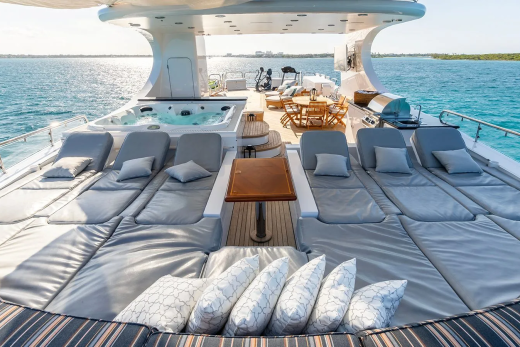 Plush lounge chairs and a hot tub fill the foreground of a luxurious yacht, while the turquoise ocean and distant coastline stretch across the background.