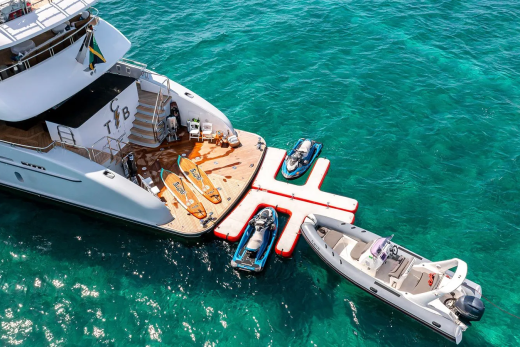 A large yacht with watercraft attached to its stern floats in clear turquoise water.