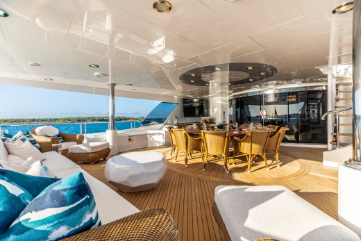 A luxurious yacht deck with comfortable seating and a dining area in the foreground, overlooking a calm blue sea and distant coastline in the background.