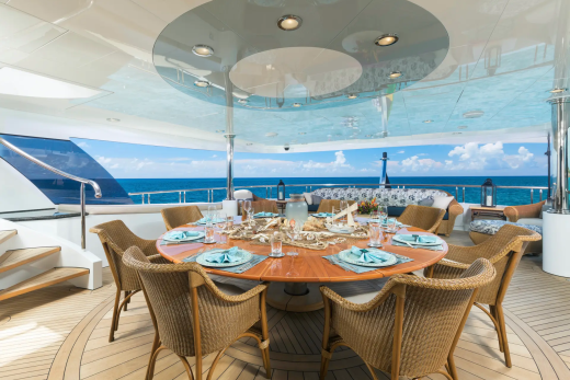 A circular dining table set for a meal on a yacht's deck is surrounded by wicker chairs, with the ocean and sky visible in the background.