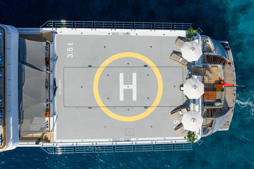 A helipad is visible on the deck of a yacht, with lounge chairs and umbrellas set up on the stern, all surrounded by clear blue water.