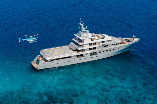 A large gray yacht with a helipad is anchored in clear blue water, with a helicopter flying nearby.