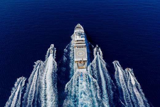 A large yacht with a helipad is trailed by several smaller boats creating white wakes in the deep blue ocean.