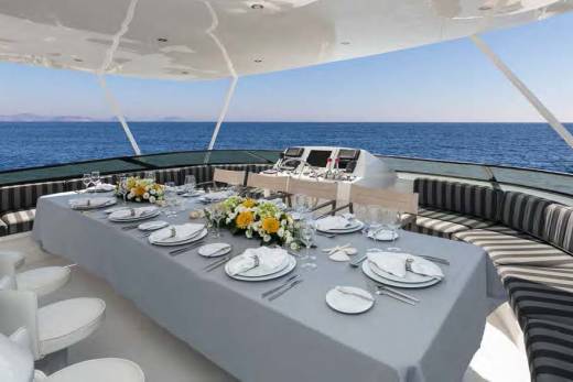 A long dining table set for a meal on a yacht's deck is in the foreground, with the vast blue ocean and clear sky in the background.