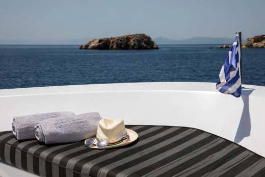 Two rolled towels, a hat, and sunglasses rest on a striped lounge chair in the foreground, with a Greek flag waving beside a rocky island and the calm blue sea in the background.