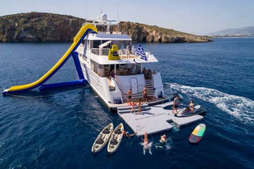 A large yacht with an inflatable slide and water toys is anchored in a clear blue bay with rocky islands in the background.