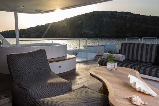 A luxurious yacht deck with a hot tub and seating area in the foreground overlooks a tranquil bay at sunset.