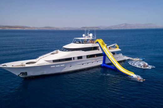 A large white yacht with a yellow inflatable slide extending into the blue ocean is anchored in front of a hazy, mountainous coastline under a clear sky.