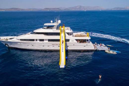 A large white yacht with a yellow slide extending into the deep blue sea is anchored in the background, while a person swims in the foreground.