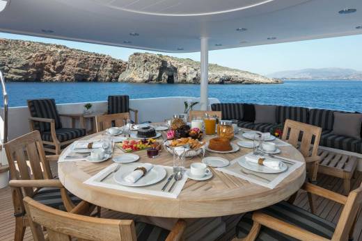 A lavish breakfast spread is set on a round wooden table on a yacht's deck, with a rocky coastline and blue sea in the background.