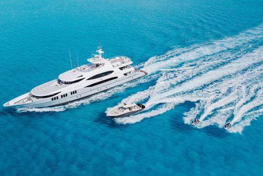 A large white yacht cruises through turquoise waters, trailed by a smaller boat and two jet skis.