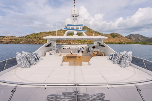 A luxurious yacht with plush seating and sunpads in the foreground is anchored in calm blue water with a lush, green, mountainous coastline in the background.
