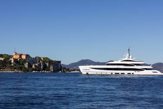A large white yacht sails on calm blue water in the foreground, with a historic stone building on a cliff and distant mountains in the background under a clear blue sky.