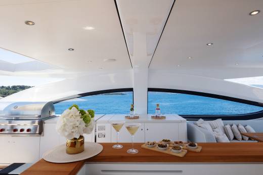A wooden bar with cocktails and appetizers in the foreground is set against a backdrop of the ocean viewed through a large window on a modern yacht.