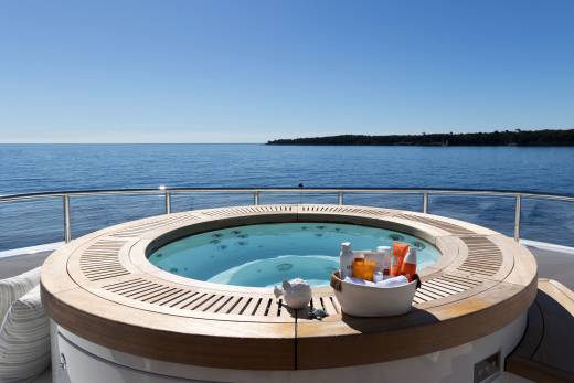 A circular jacuzzi with toiletries sits on a yacht's deck, overlooking a calm blue sea and a distant, tree-lined coast under a clear sky.