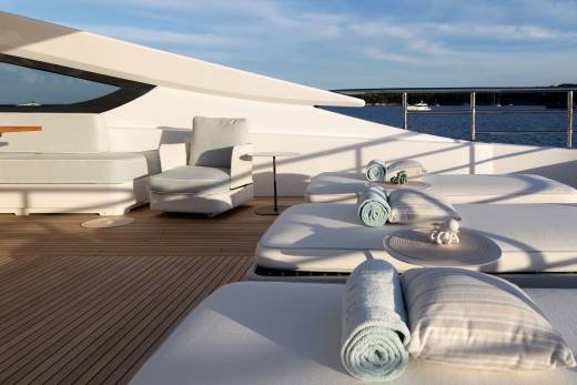 Rolled towels and lounge chairs on a yacht's deck are in the foreground, with a calm body of water and a distant bridge in the background.