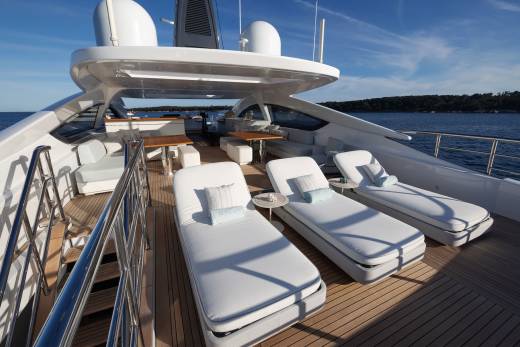 Three white lounge chairs with blue pillows sit on the wooden deck of a modern yacht in the foreground, with a calm blue ocean and a clear sky in the background.