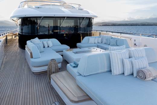 A luxurious yacht's deck, furnished with plush light blue seating and a fire pit, overlooks a calm sea and distant, hazy coastline.