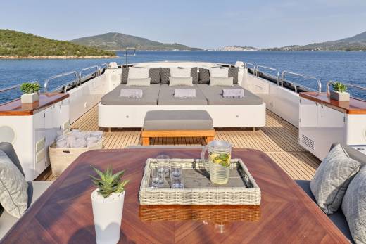 A luxurious yacht deck with comfortable seating and a refreshing drink in the foreground is anchored in a calm, blue sea with lush green islands in the background.