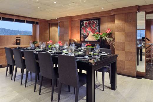 A formal dining table set for a large group is in the foreground, while a hallway with a staircase and a glimpse of the outdoors are in the background.