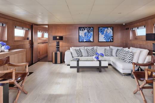 Two wooden chairs with striped side tables are in the foreground, while a large white sectional sofa with blue and white patterned pillows sits in the background, facing two framed blue floral artworks on a wood-paneled wall.
