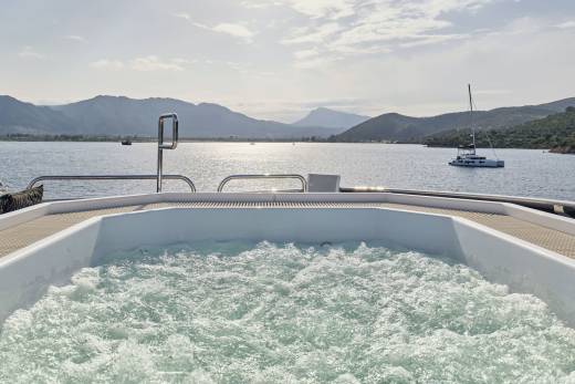 A bubbling hot tub on a yacht's deck overlooks a calm bay with distant, hazy mountains and a sailboat.