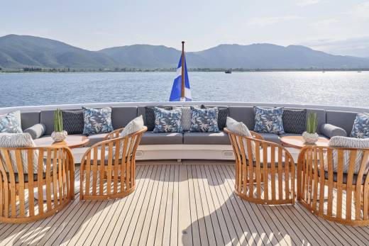Wooden chairs and plush seating arrangements on a yacht's deck face a calm blue sea and distant, hazy mountains under a bright sky.