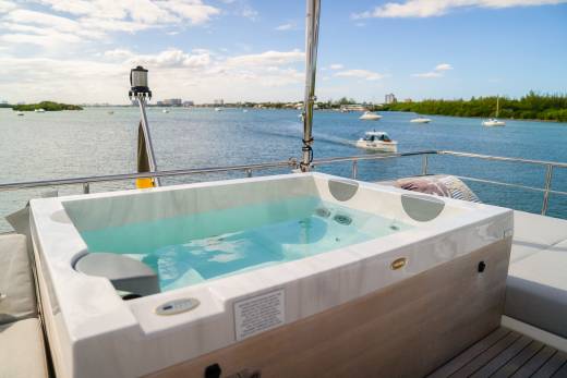 A luxurious hot tub filled with clear blue water sits on a yacht's deck, with a calm bay dotted with boats and lush green islands in the background.