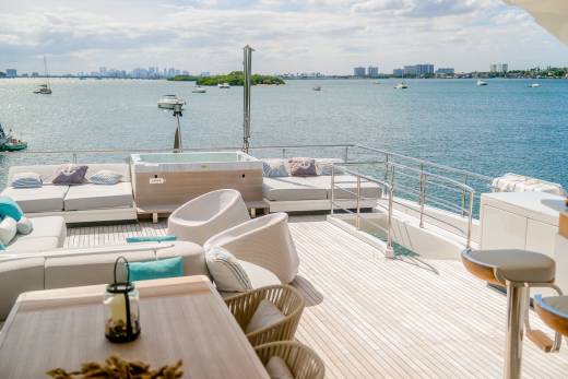 A luxurious yacht deck with comfortable seating and a hot tub in the foreground overlooks a city skyline across a calm bay.