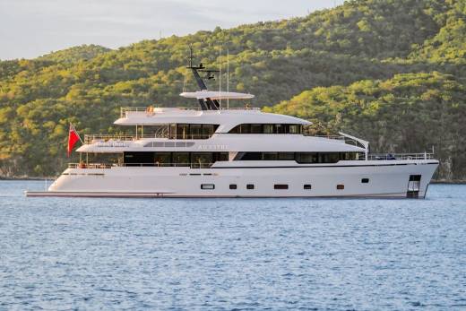 A large white yacht sails on calm blue water in the foreground, with lush green hills rising in the background.