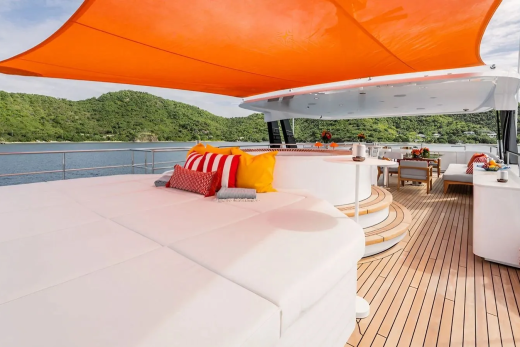 A spacious white sunbed with colorful pillows sits on a wooden deck under an orange canopy, with a jacuzzi and dining area visible, all set against a backdrop of lush green hills and calm blue water.