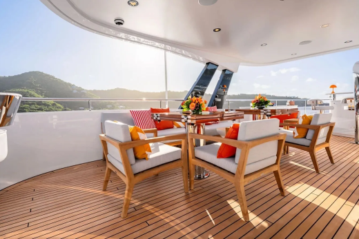 A sun-drenched yacht deck with comfortable seating and a dining table in the foreground is set against a backdrop of lush green hills under a clear blue sky.