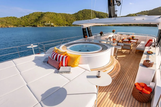 A luxurious yacht with a hot tub and lounge area in the foreground is anchored in a calm bay with lush green hills in the background.