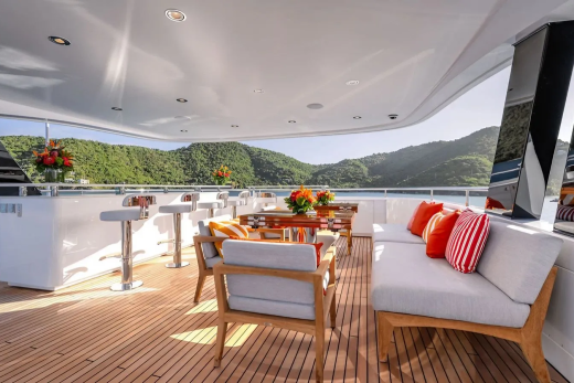 A luxurious yacht deck with comfortable seating and a bar in the foreground overlooks a lush, green coastline under a bright sky.