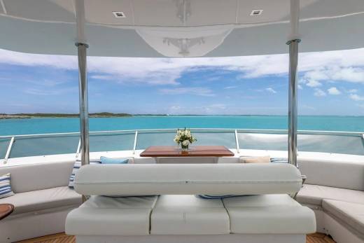 A white cushioned seating area with a wooden table holding a floral arrangement is in the foreground, overlooking a calm turquoise ocean under a bright blue sky in the background.
