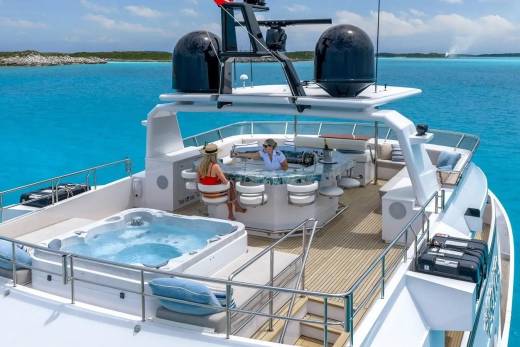 A luxurious yacht with a hot tub in the foreground cruises through clear turquoise waters with a distant, low-lying island in the background.