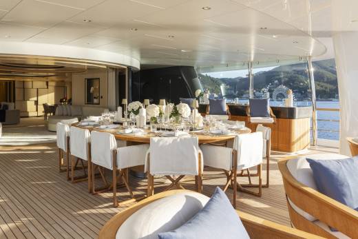 A luxurious yacht deck features a beautifully set dining table in the foreground, with comfortable seating and a scenic view of a sun-drenched coastline in the background.