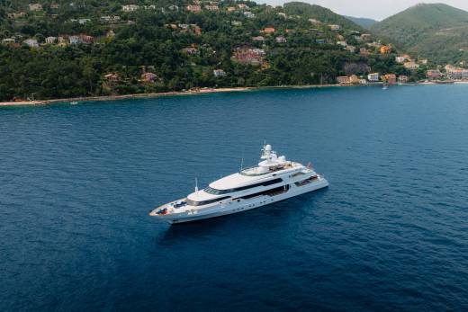 A large white yacht floats on calm blue water in the foreground, with a lush green, hilly coastline dotted with houses in the background.