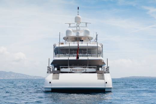 A large white yacht with the name "SHAI LUXURY" on its stern is at sea with a hazy coastline in the background under a cloudy sky.