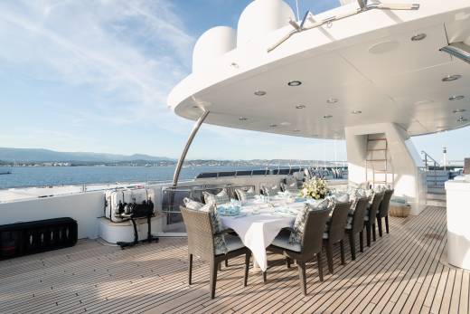 A long dining table set for a meal on a yacht's deck overlooks a calm sea and distant coastline under a bright sky.